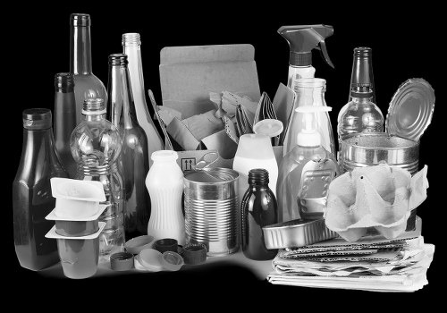 A collection of various glass bottles, plastic containers, and paper packaging arranged on a surface, with some bottles standing upright and others leaning or stacked. The glass bottles include clear and dark-colored types with different shapes and cap styles, while the plastic containers vary in size and texture, featuring spray bottles, jars, and cups. There is a pile of crumpled paper or newspaper situated on the right side of the image, alongside metallic cans and small miscellaneous items. The background appears dark or plain, highlighting the assortment of rubbish materials, which are likely prepared for removal or disposal. This visual is relevant to rubbish removal services, such as those offered by Commercial Waste Removal Sidcup, operating in the local area, including Sidcup postcodes. The scene depicts typical items cleared during commercial waste collection, emphasizing the need for professional rubbish removal in the region.