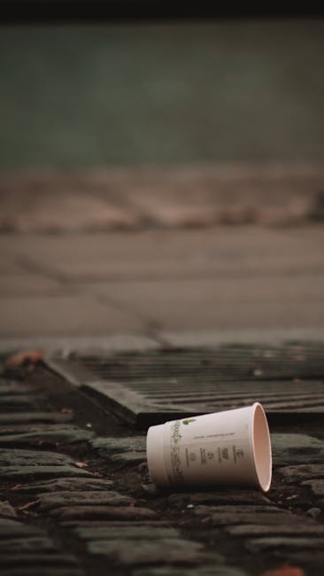 Close-up photograph of a tightly gathered, large black plastic rubbish bag with visible creases and a glossy surface, positioned in the foreground. The bag is placed on a solid surface against a plain