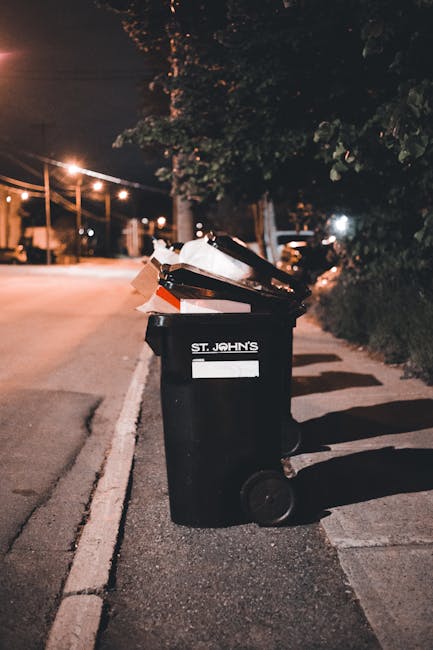 Two large black plastic trash bags containing waste are placed on a paved sidewalk beside a black metal fence with vertical bars. The bags are crumpled and appear to be filled with household or garden