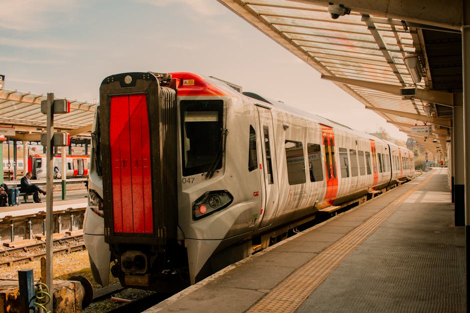 A modern electric train with a sleek silver body and red accents is stationary at an outdoor train station platform. The train's front section features a black and red protective cover, and its smooth